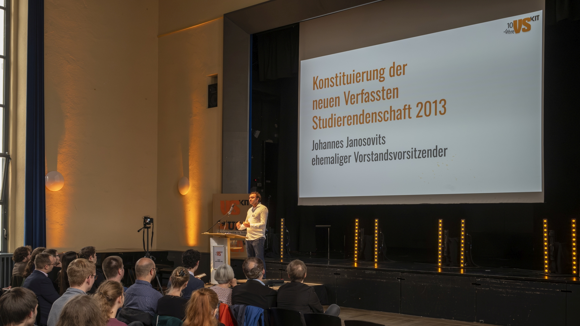 Johannes Jonosovits hält eine Rede beim 10-jähigen Jubiläum der Verfassten Studierendenschaft am KIT. Im Hintergrund ist eine Vortragsfolie, auf der steht: Konstituierung der neuen Verfassten Studierendenschaft 2013.