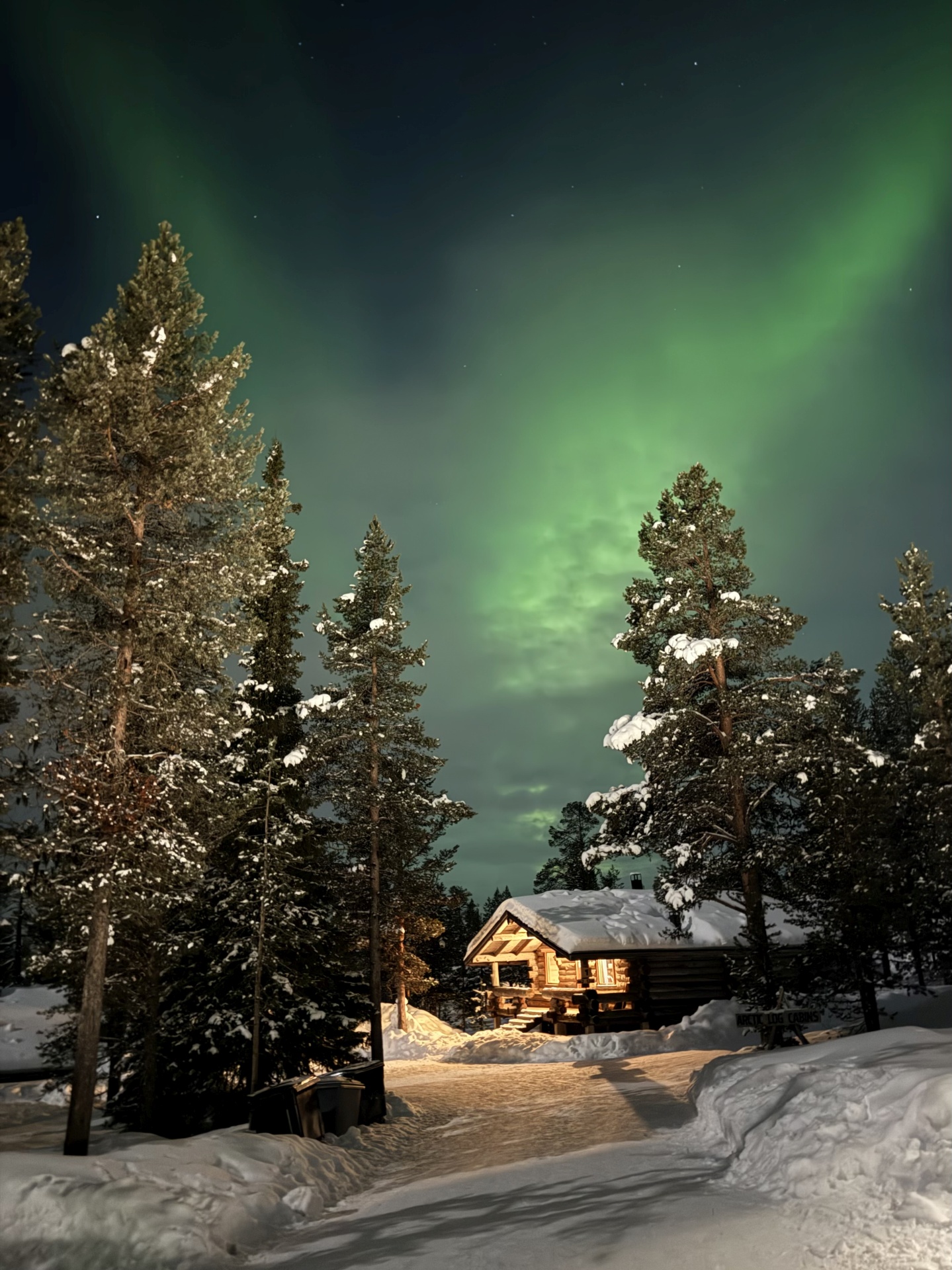 Verschneite Hütte im Wald unter Polarlichtern in der Nacht.