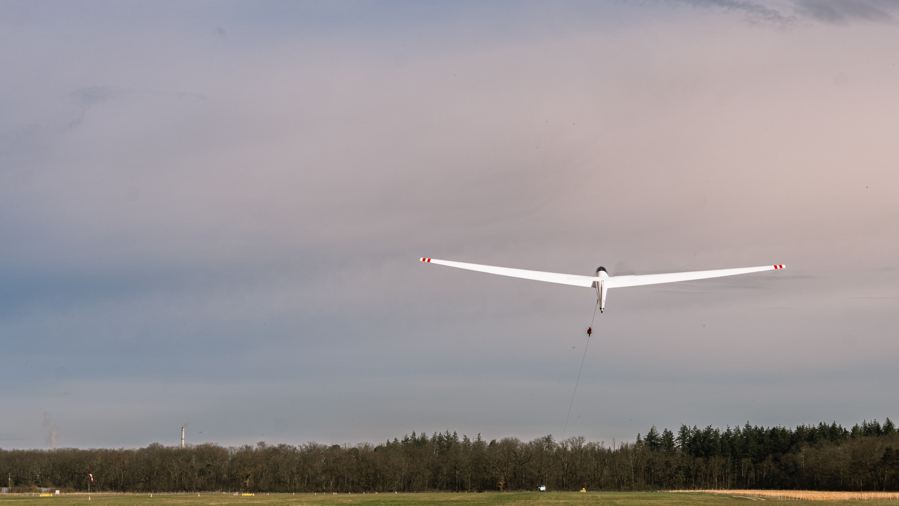 Glider aircraft mid-air over field, hanging from a cable with a rope and trees in the background.