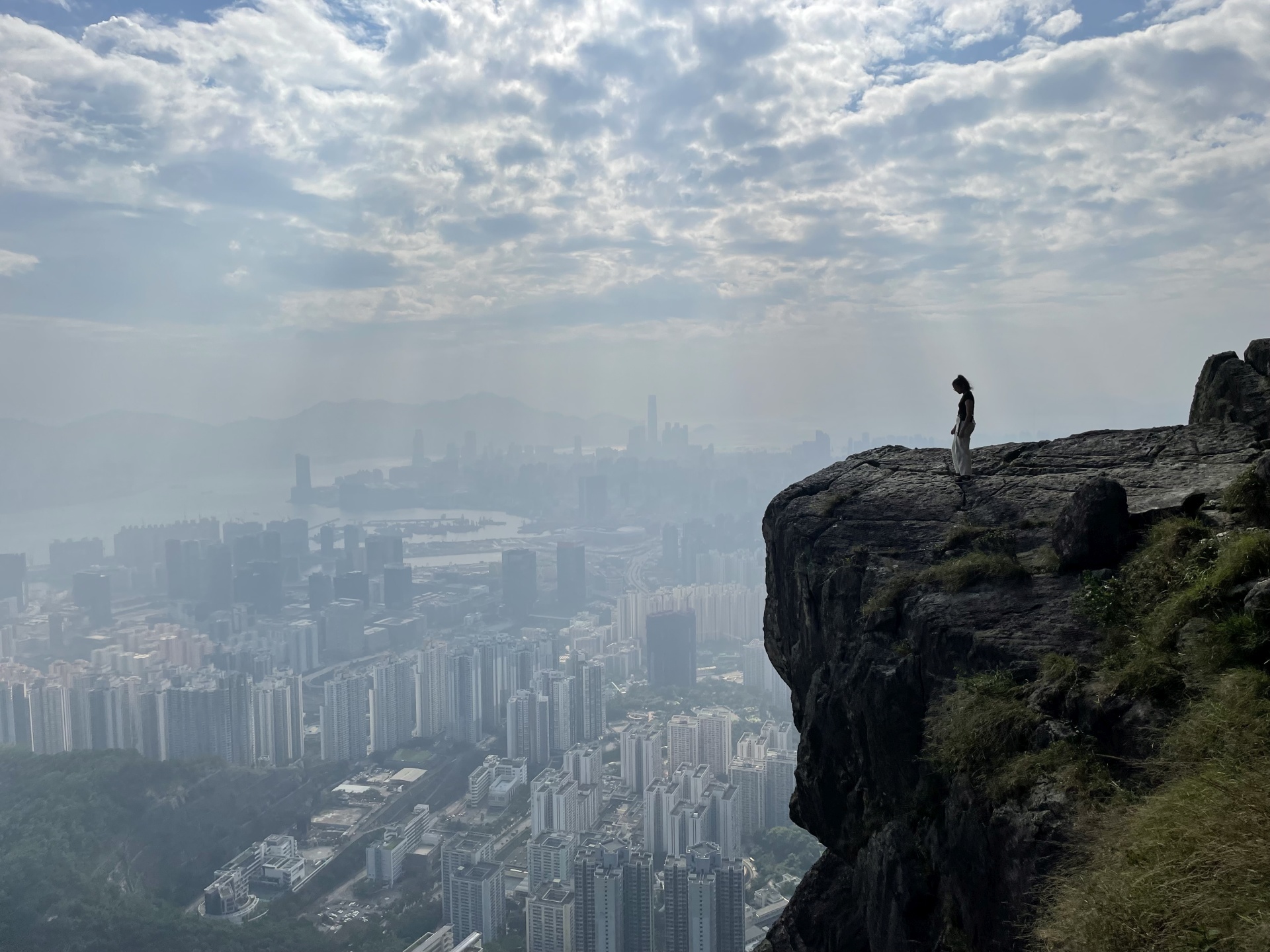 Person steht auf einem Felsen mit Blick über eine Stadt bei wolkigem Himmel.