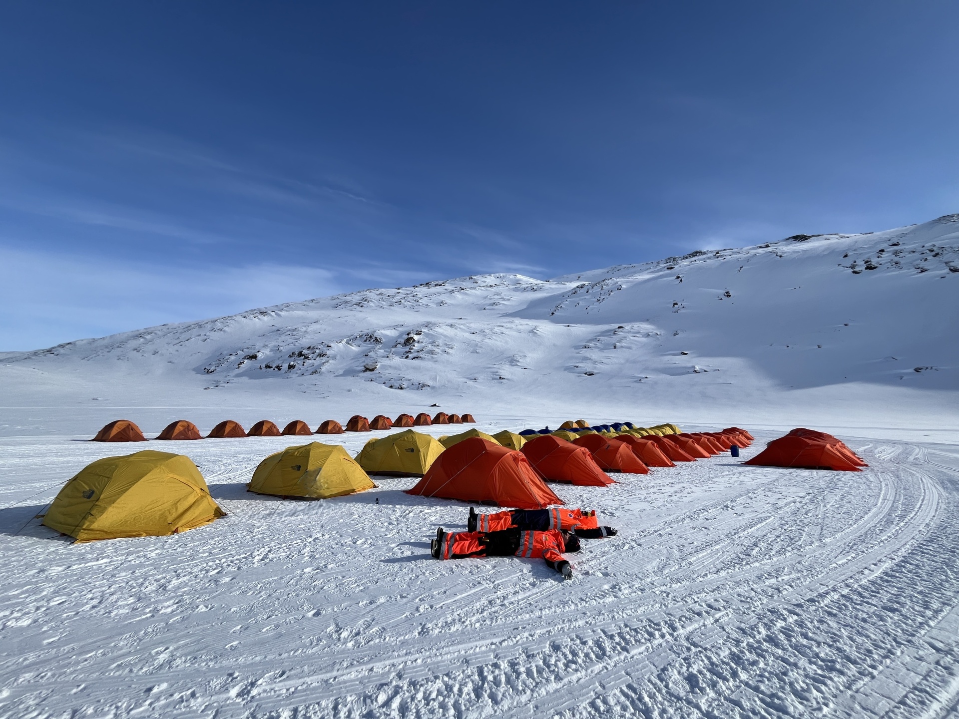 Zelte im Schnee vor verschneiter Berglandschaft unter klarem Himmel.