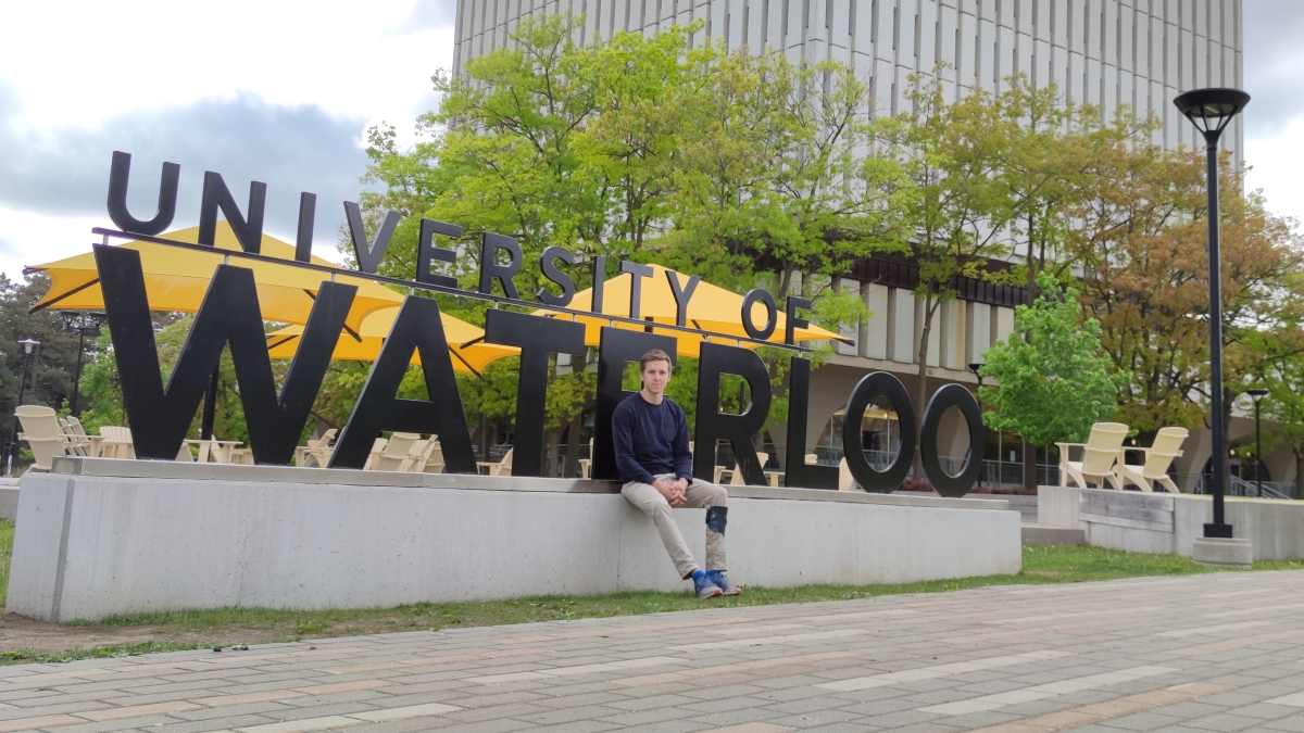 Kristof sitzt vor dem Schild, auf dem steht University of Waterloo. 