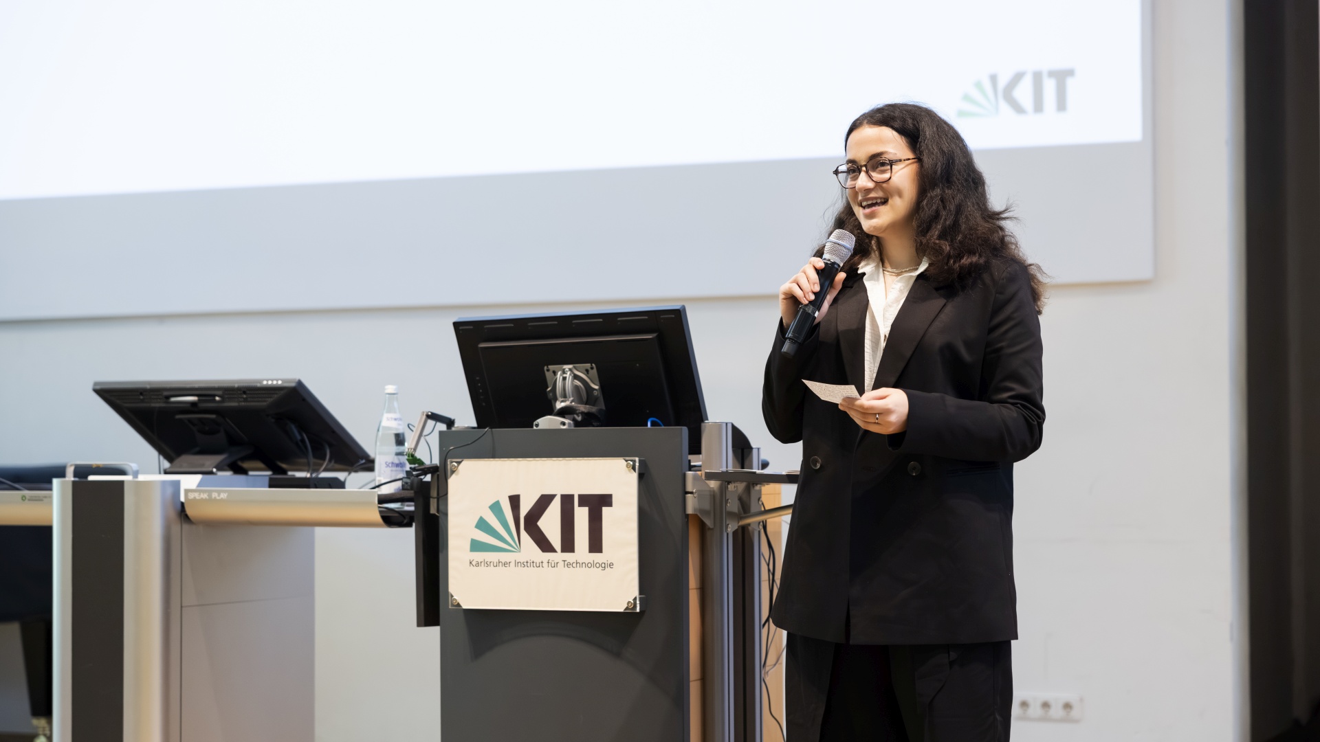 Defne Çelik stands on a stage with a microphone and some cards in her hands. 