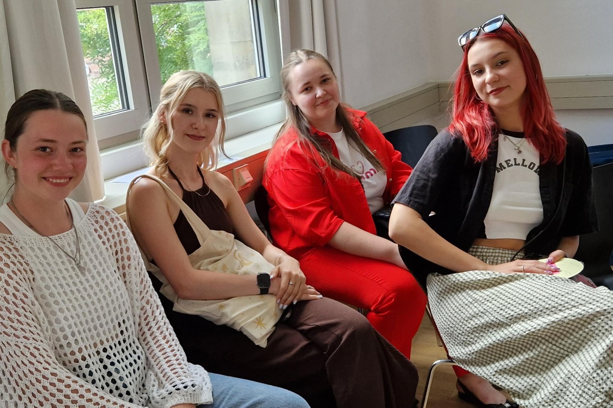 Vier junge Frauen sitzen nebeneinander und blicken l&auml;chelnd in die Kamera.