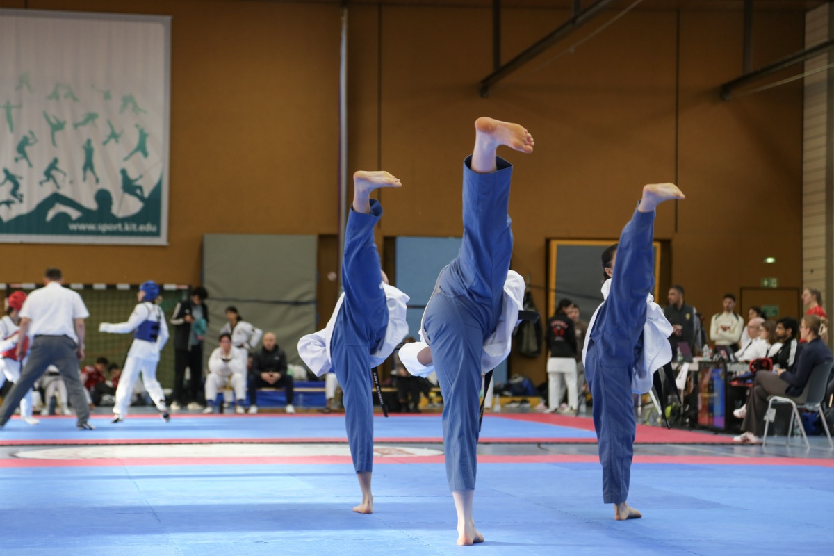 Drei Taekwondo-Kämpfer führen im Wettkampf auf der Matte Vorwärtskicks aus; Zuschauer im Hintergrund.