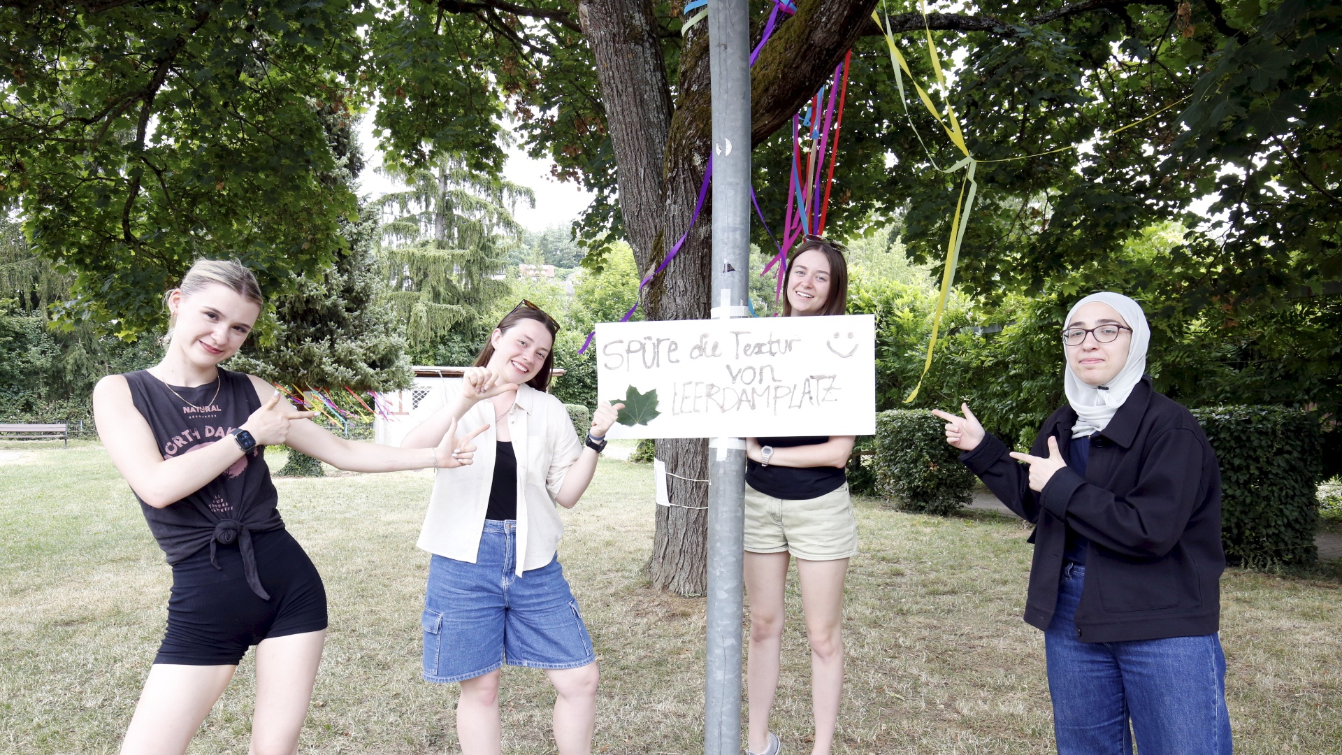 Four young women standing outdoors next to a sign that reads "Spüre die Textur von Leerdamplatz".