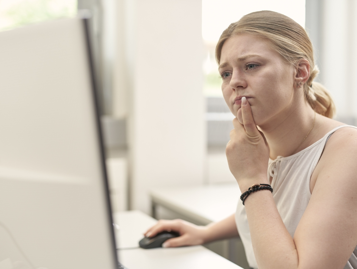 Eine Frau sitzt mit fragendem Gesicht und Zeigefinger vor dem Mund, vor ihrem Bildschirm. 