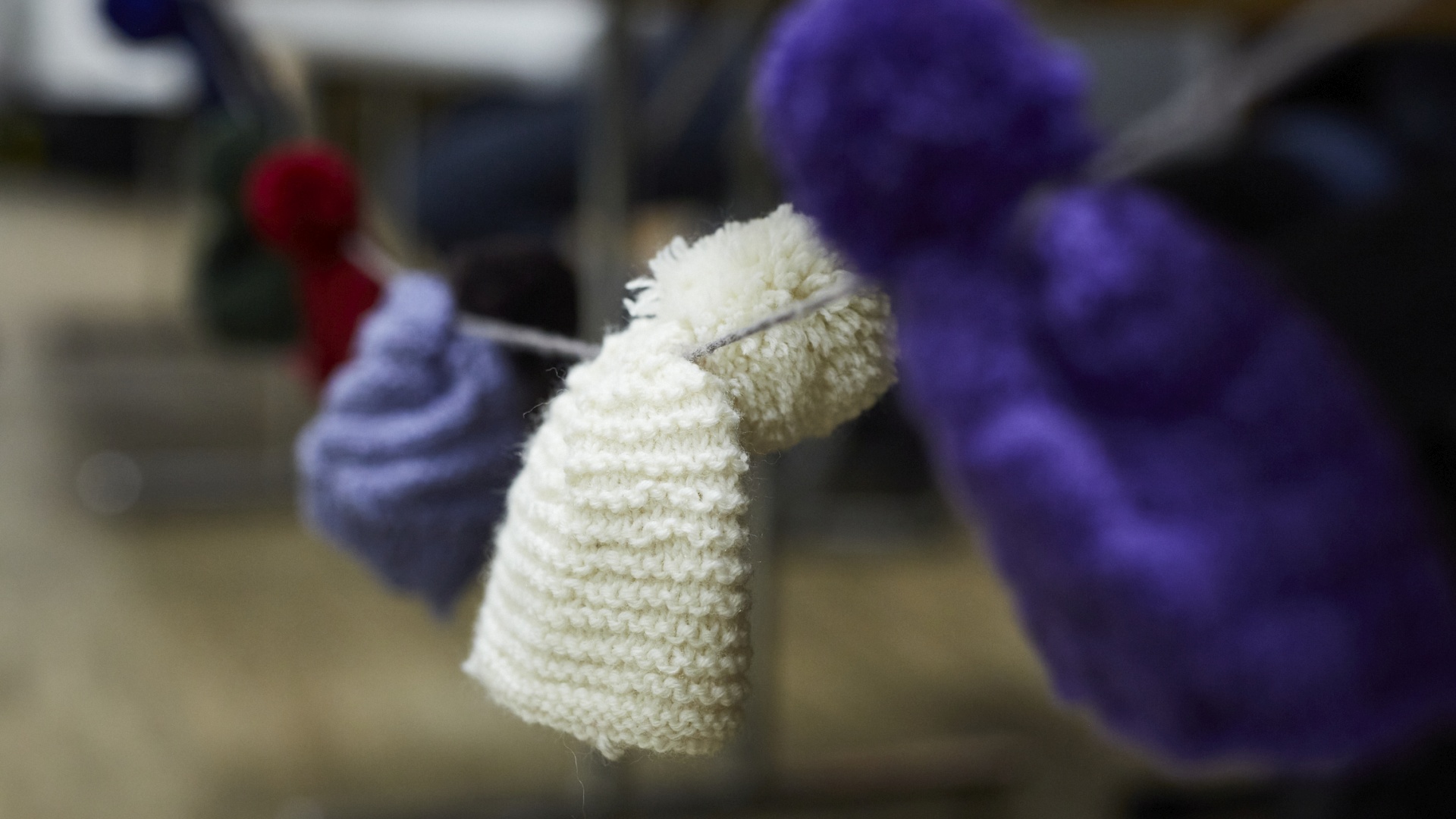 Weiß gestrickter Mini-Artikel hängt an einer Schnur, bunte Wollkugeln im Hintergrund.